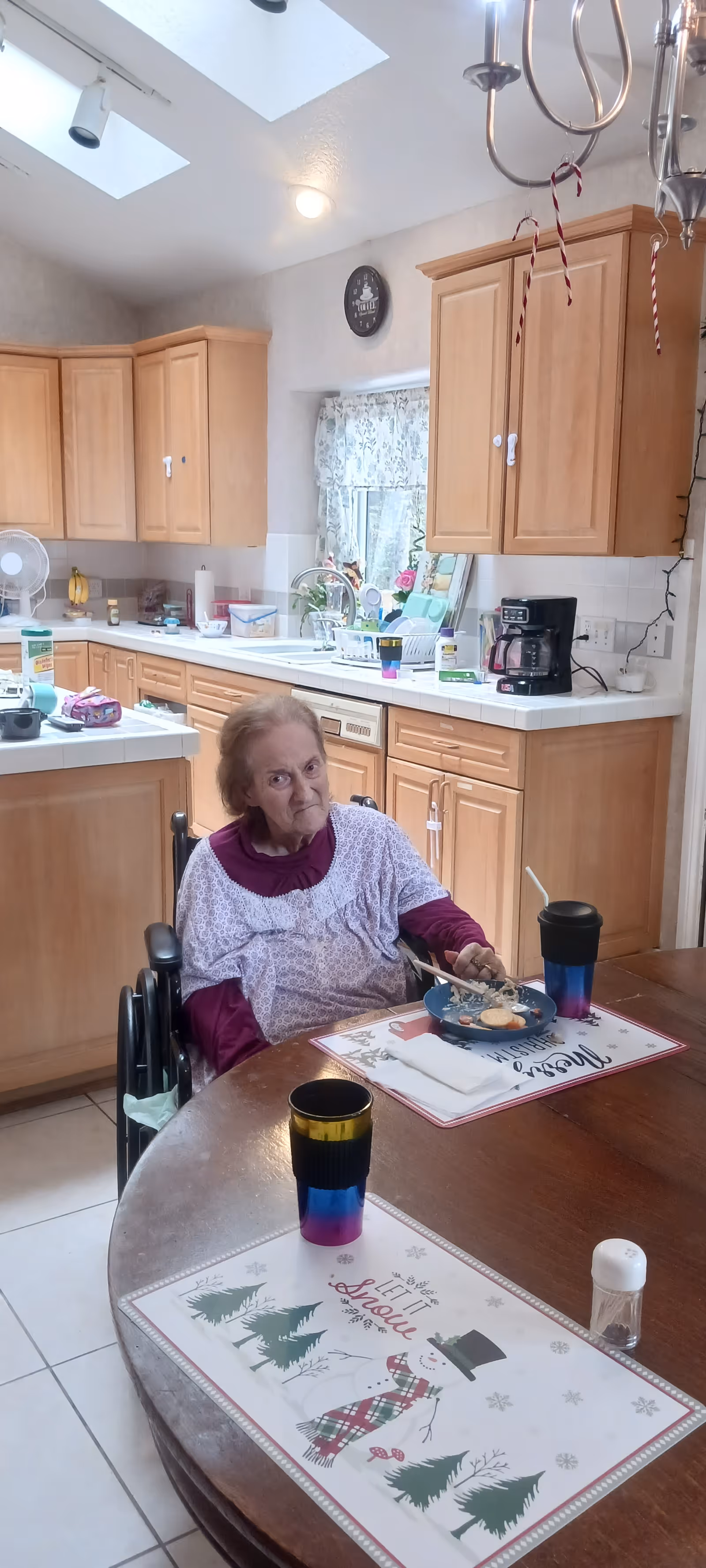 An elderly woman in a wheelchair eating at a round wooden dining table in a kitchen. The kitchen has light wood cabinets, a coffee maker, and various kitchen items on the counters. The table has placemats with winter and holiday designs, two colorful cups, and a salt shaker. The room is well-lit with natural light from skylights and a window with floral curtains.