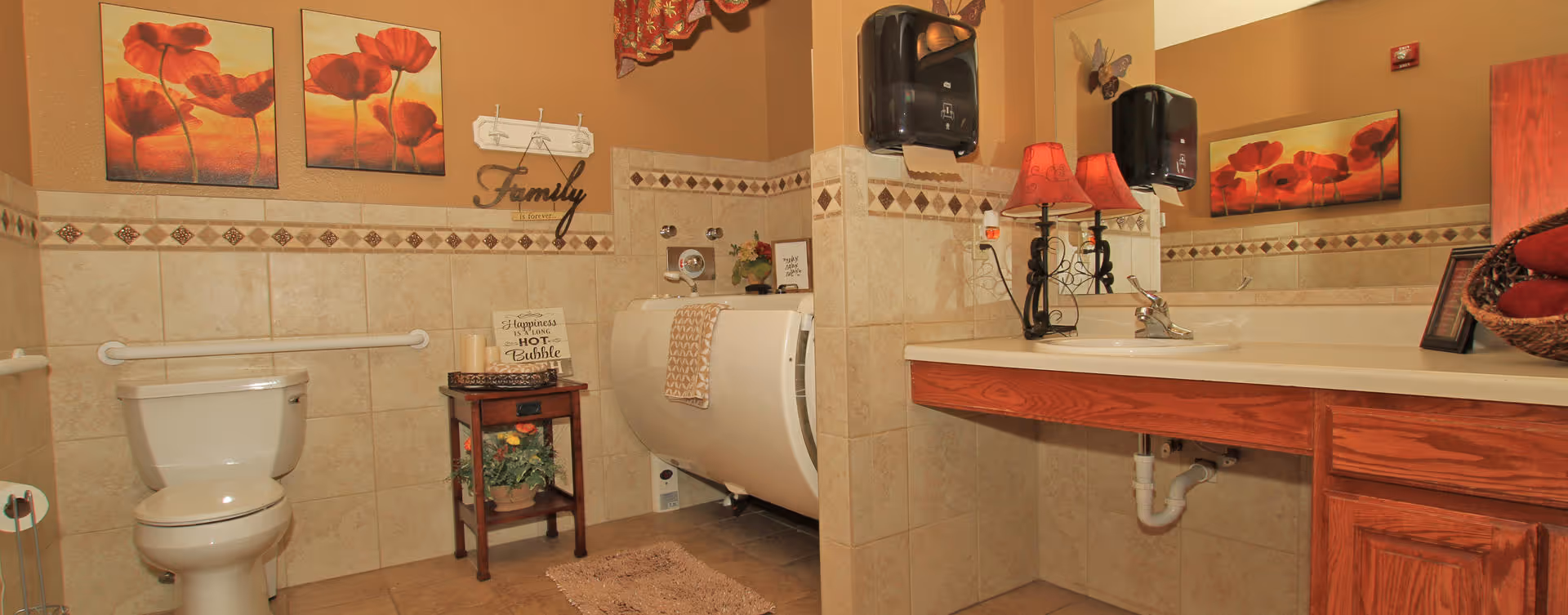 A bathroom with beige tiled walls and floor, featuring a white toilet, a walk-in bathtub with a towel draped over the side, a wooden side table with decorative items, a large mirror above a wooden vanity with a sink, two red lamps on the vanity, and wall art with red flowers and the word 'Family'.