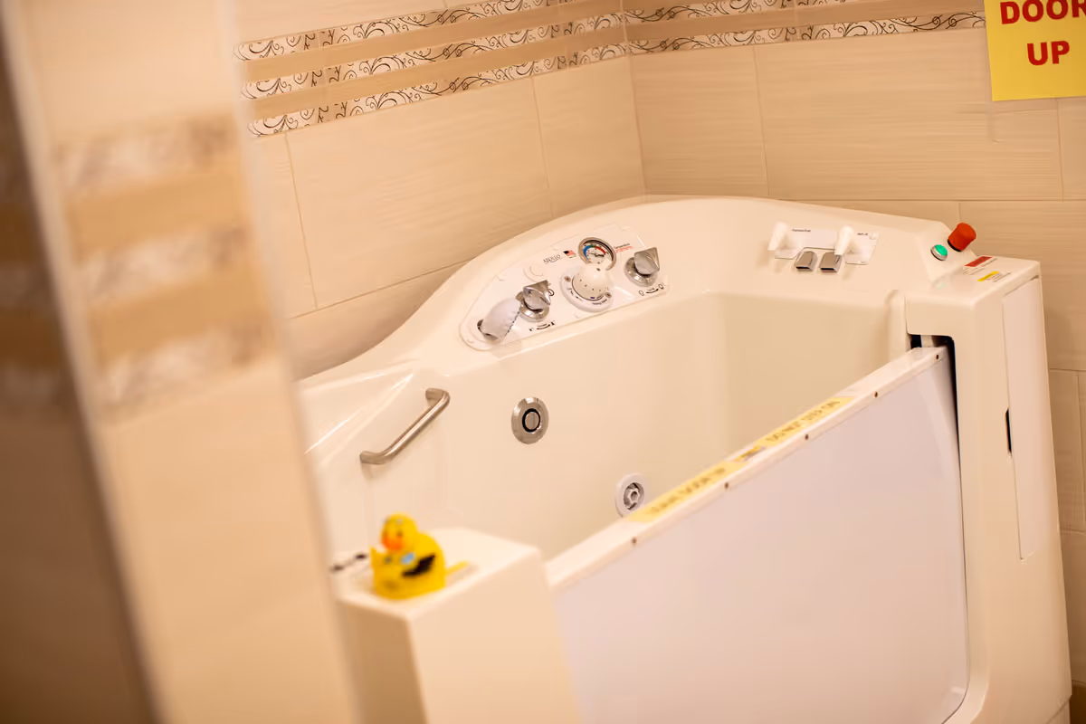 A white walk-in bathtub with control knobs, a safety handle, and a rubber duck in a tiled bathroom.