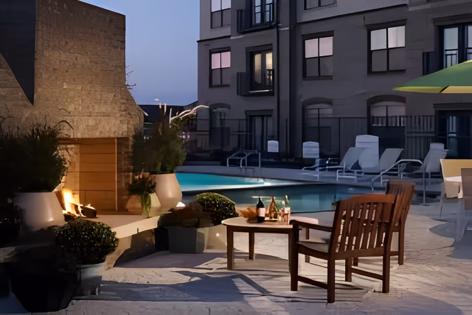Outdoor patio area at dusk with a lit stone fireplace, wooden chairs around a small table with bottles and glasses, potted plants, and a swimming pool in the background surrounded by lounge chairs and a building.