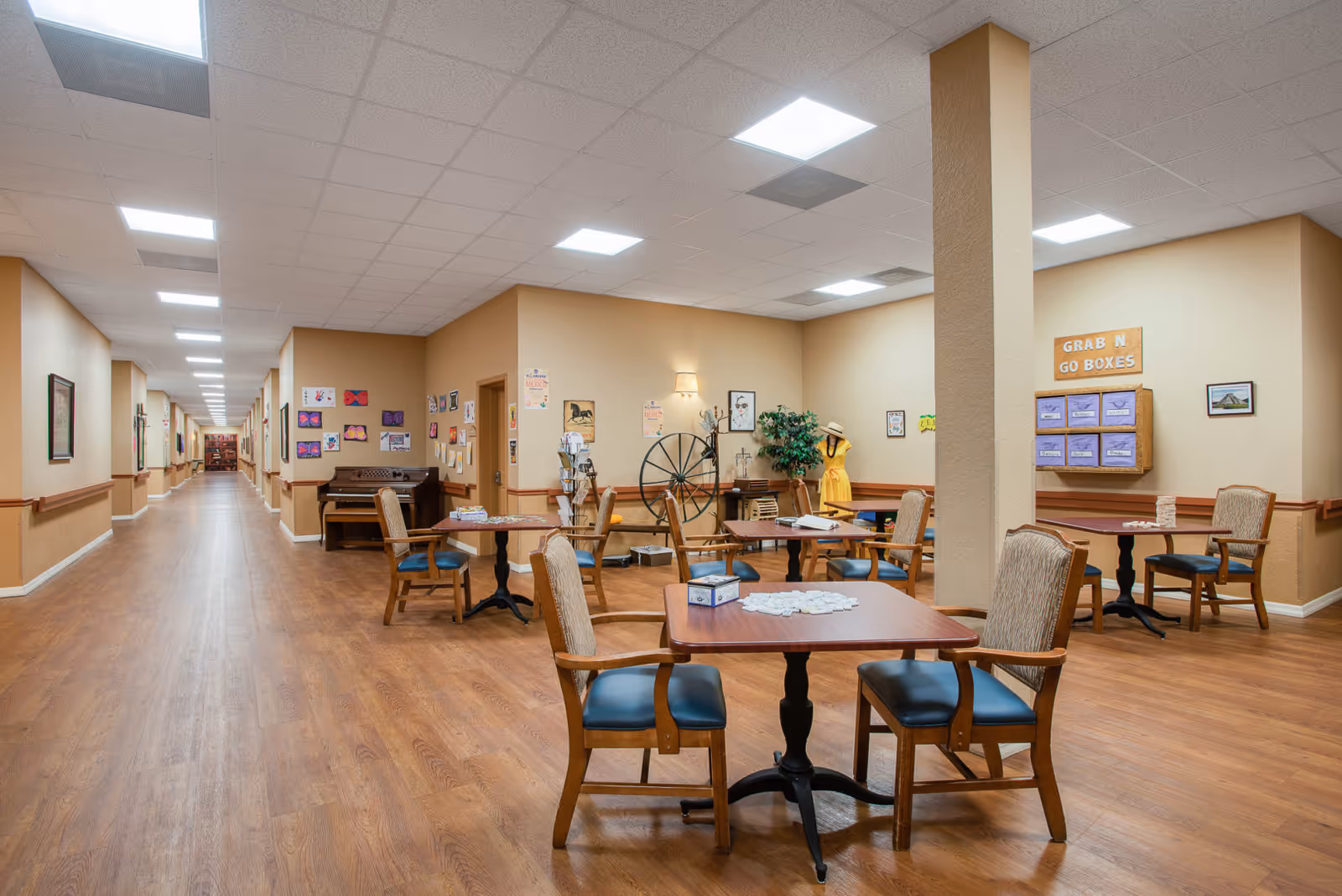 A spacious common area in a senior living facility with several wooden tables and chairs arranged for games and activities. The room has wood flooring, beige walls decorated with artwork and posters, a piano against the wall, and a display labeled 'Grab N Go Boxes'. A long hallway extends from the room with more artwork on the walls.
