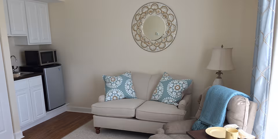 A cozy living area with a beige loveseat adorned with two decorative blue and white patterned pillows. To the right is a beige armchair with a blue throw blanket draped over it and a side table with a lamp. On the left side, there is a small kitchenette with white cabinets, a sink, a microwave, and a mini refrigerator. A decorative round mirror hangs on the wall above the loveseat. The room has wooden flooring and light-colored walls with a window covered by blue curtains on the right.