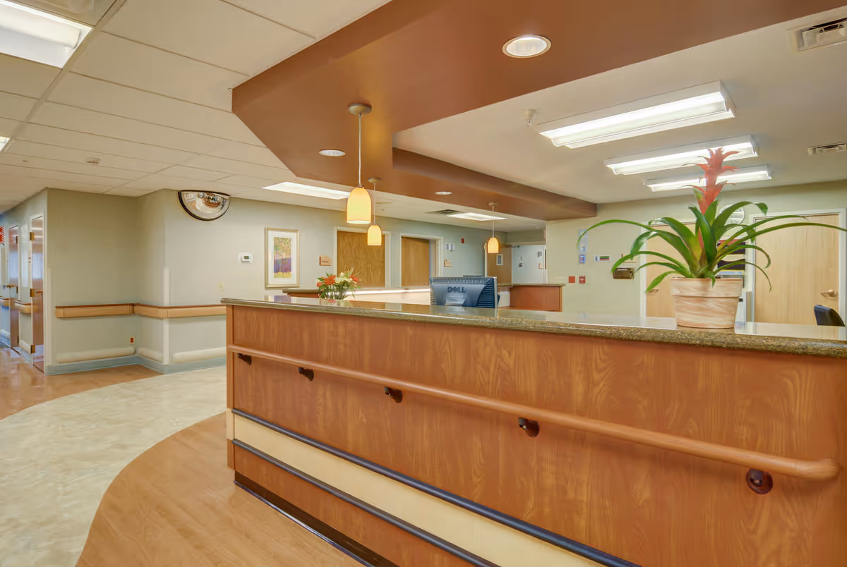 Reception desk area inside a senior living facility with a wooden counter, a computer monitor, hanging pendant lights, a potted plant, and a hallway leading to rooms in the background.