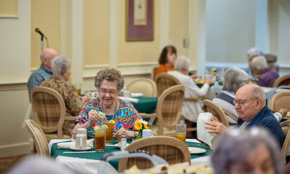 A group of elderly people sitting at tables in a dining room, enjoying their meals and drinks. The room has beige walls and framed artwork, with green tablecloths on the tables. The atmosphere appears calm and social.