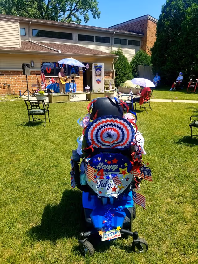 Outdoor scene at The Pillars at Crystal Bay showing a decorated mobility scooter with red, white, and blue 4th of July decorations in the foreground. In the background, a person is playing guitar under a white umbrella on a small stage attached to a brick and siding building. Several people are seated on chairs and benches on the grass, enjoying the event on a sunny day.