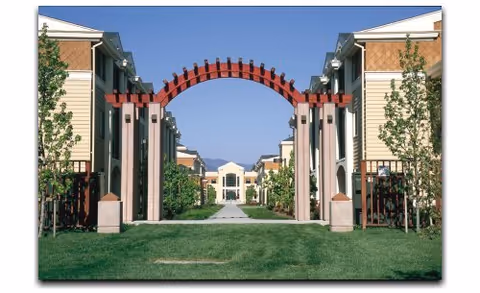 View of a residential facility with a large red wooden archway in the foreground, flanked by two beige buildings with brown shingle accents. A pathway runs through the green lawn leading to a central building in the background under a clear blue sky.