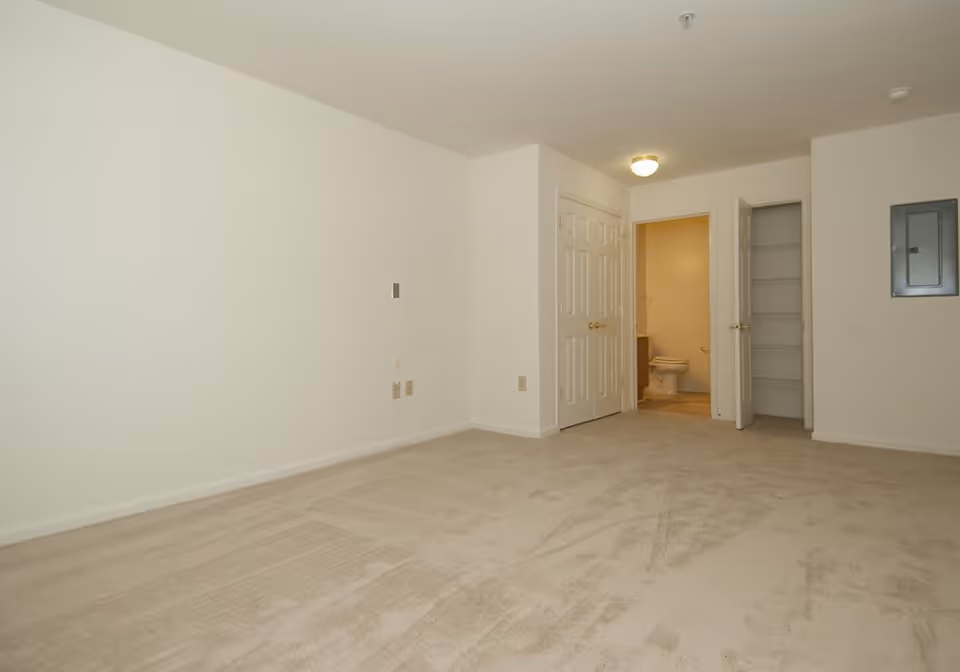 Empty carpeted room with white walls, double closet doors and an open doorway revealing a bathroom toilet.