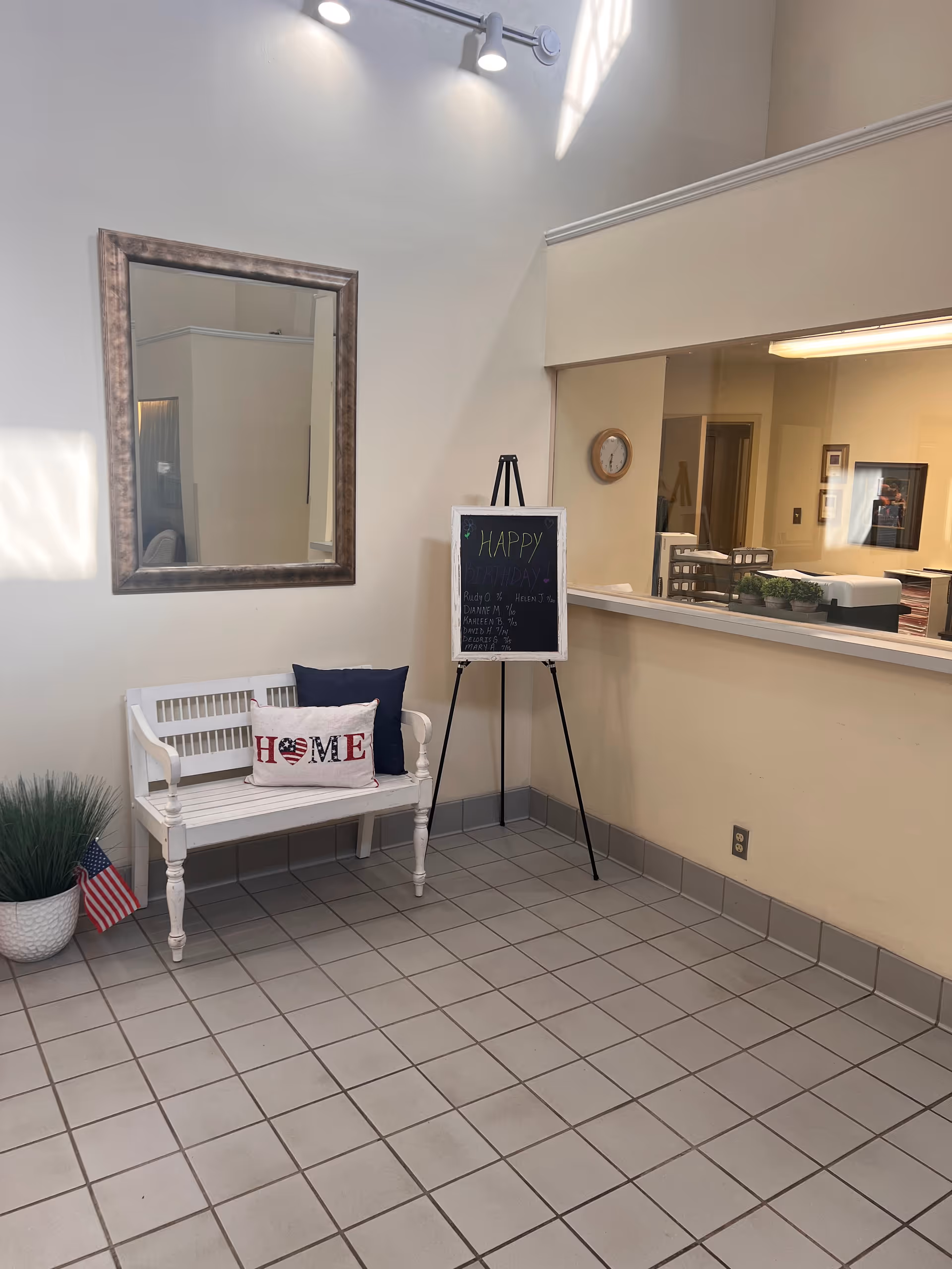 A reception area with a white bench topped with decorative pillows, an easel chalkboard, a mirror, and a service window.