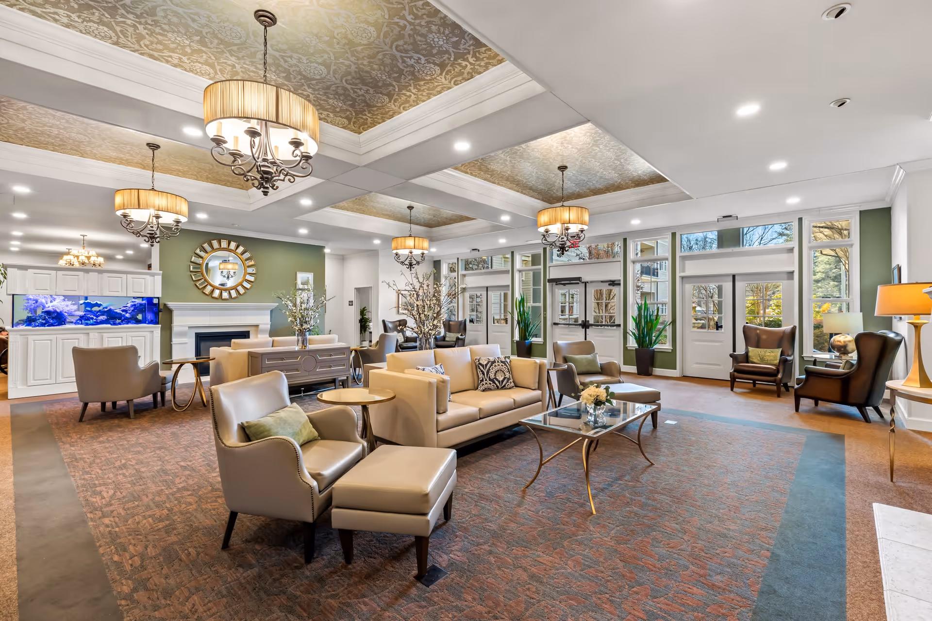 A spacious and elegantly decorated senior living facility common area with comfortable beige and brown armchairs and sofas arranged around glass coffee tables. The room features a patterned carpet, large windows letting in natural light, green accent walls, a fireplace with a decorative mirror above it, and multiple chandeliers hanging from a coffered ceiling with gold patterned insets. There is also a large aquarium on the left side and several potted plants near the windows.