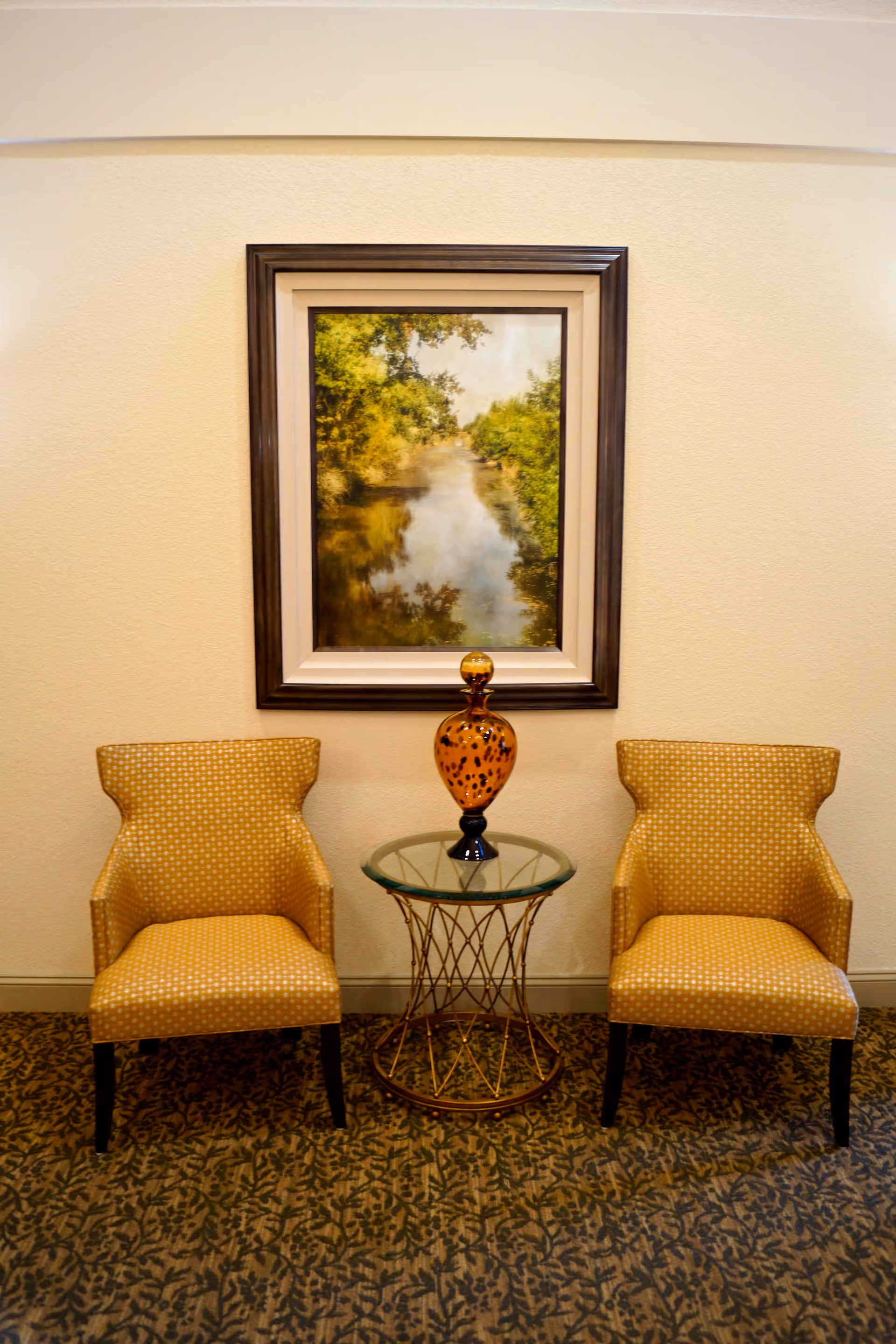 Two yellow patterned armchairs with black legs are positioned on either side of a small round glass-top table with a decorative orange vase. Behind the chairs is a framed painting of a serene river scene with trees and reflections in the water, hanging on a beige wall. The floor is covered with a patterned carpet.