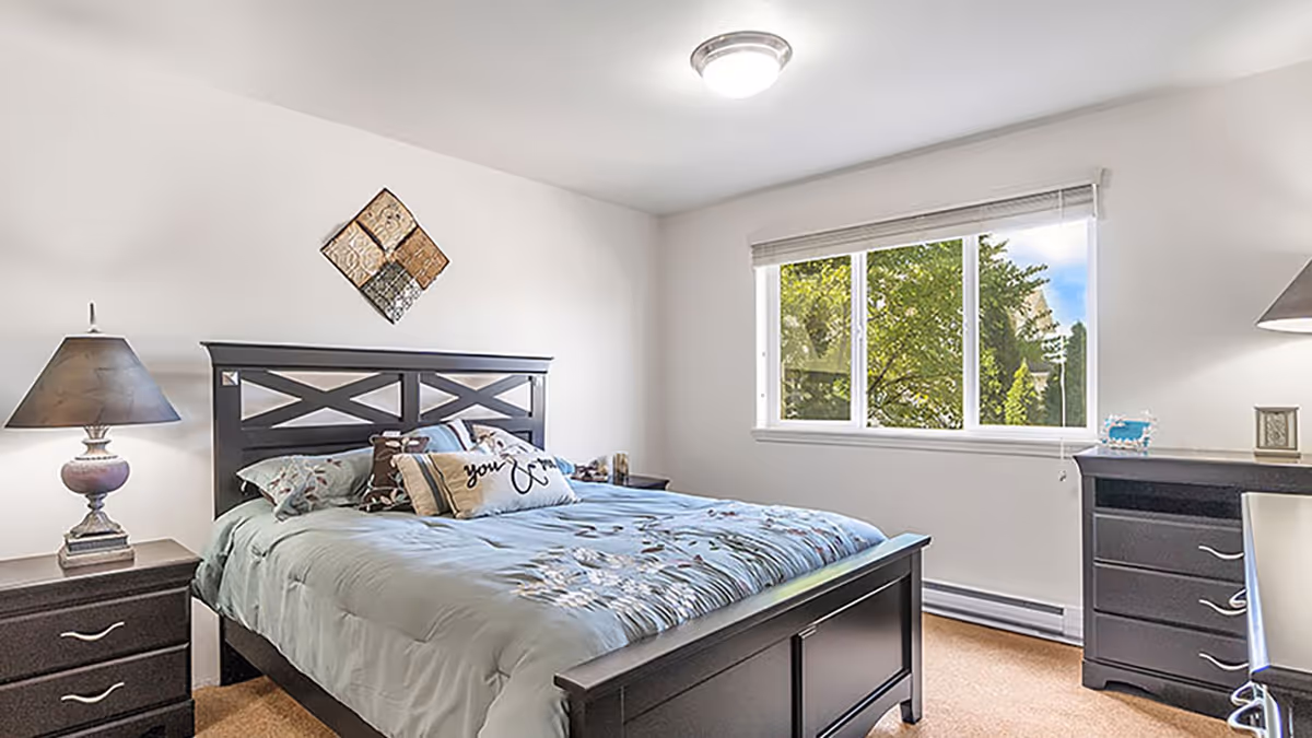 Bright bedroom with a large bed, matching nightstands and lamps, a dresser, and a window showing trees outside.