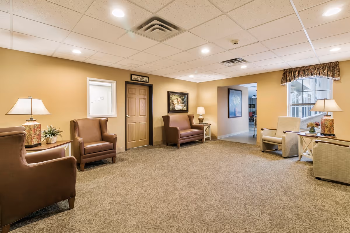 A well-lit common lounge with upholstered chairs, side tables and lamps in a carpeted senior living facility.