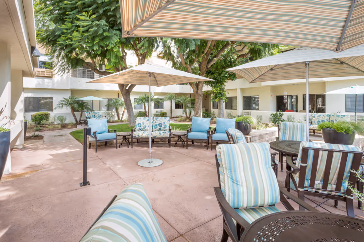 Outdoor patio area at The Groves At Tustin Assisted Living with multiple seating arrangements including cushioned chairs and sofas under large striped umbrellas, surrounded by trees and greenery.