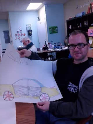 A man wearing glasses and a dark jacket is sitting indoors holding up a large drawing of a car. In the background, an older man is seated at a table with various items on it. The room has light blue walls and shelves with decorations and supplies.