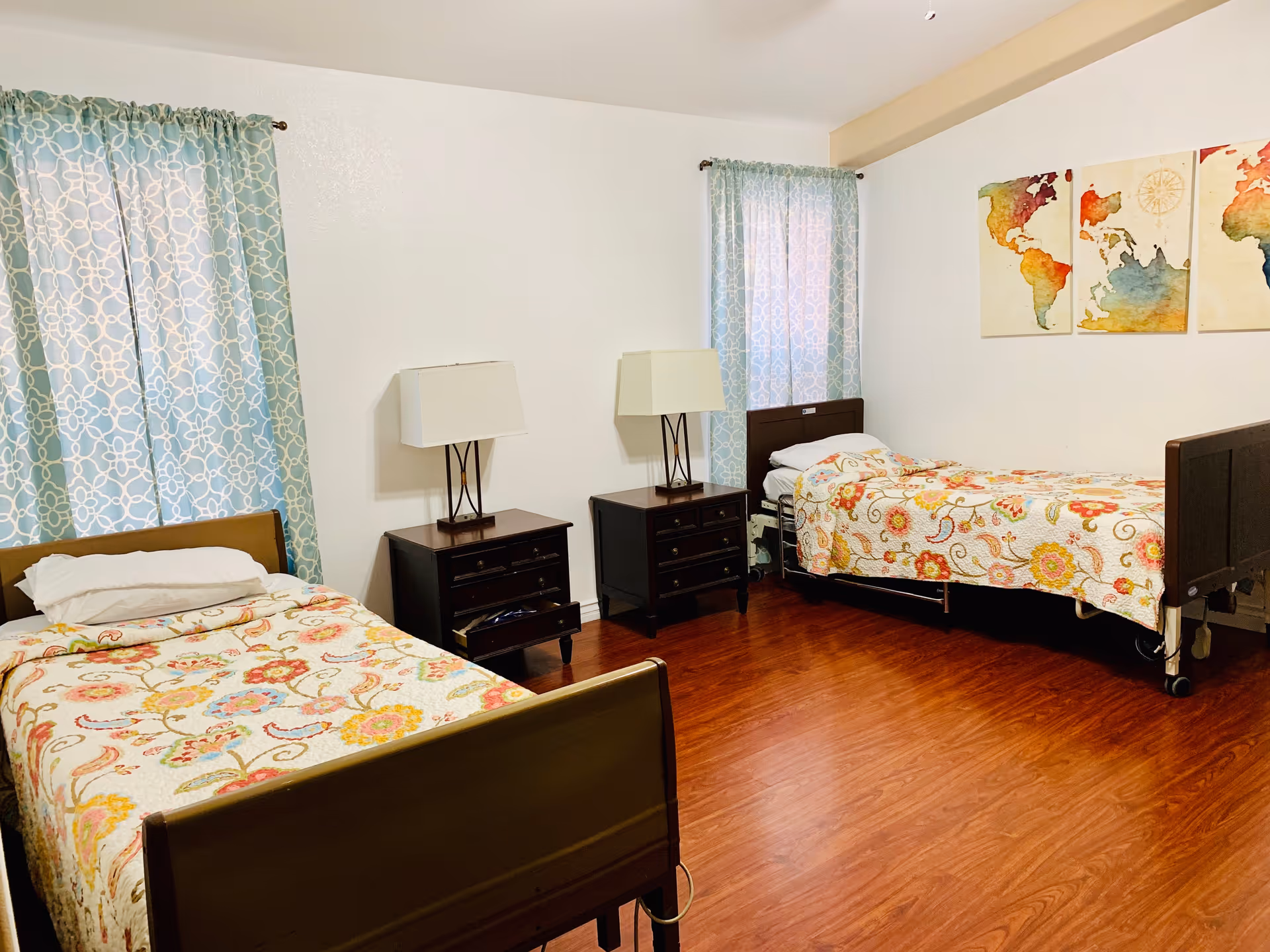 A bedroom with two single beds, each with a floral patterned bedspread. Between the beds are two dark wooden nightstands, each with a lamp. The room has wooden flooring, two windows with blue patterned curtains, and three colorful world map paintings on the wall.