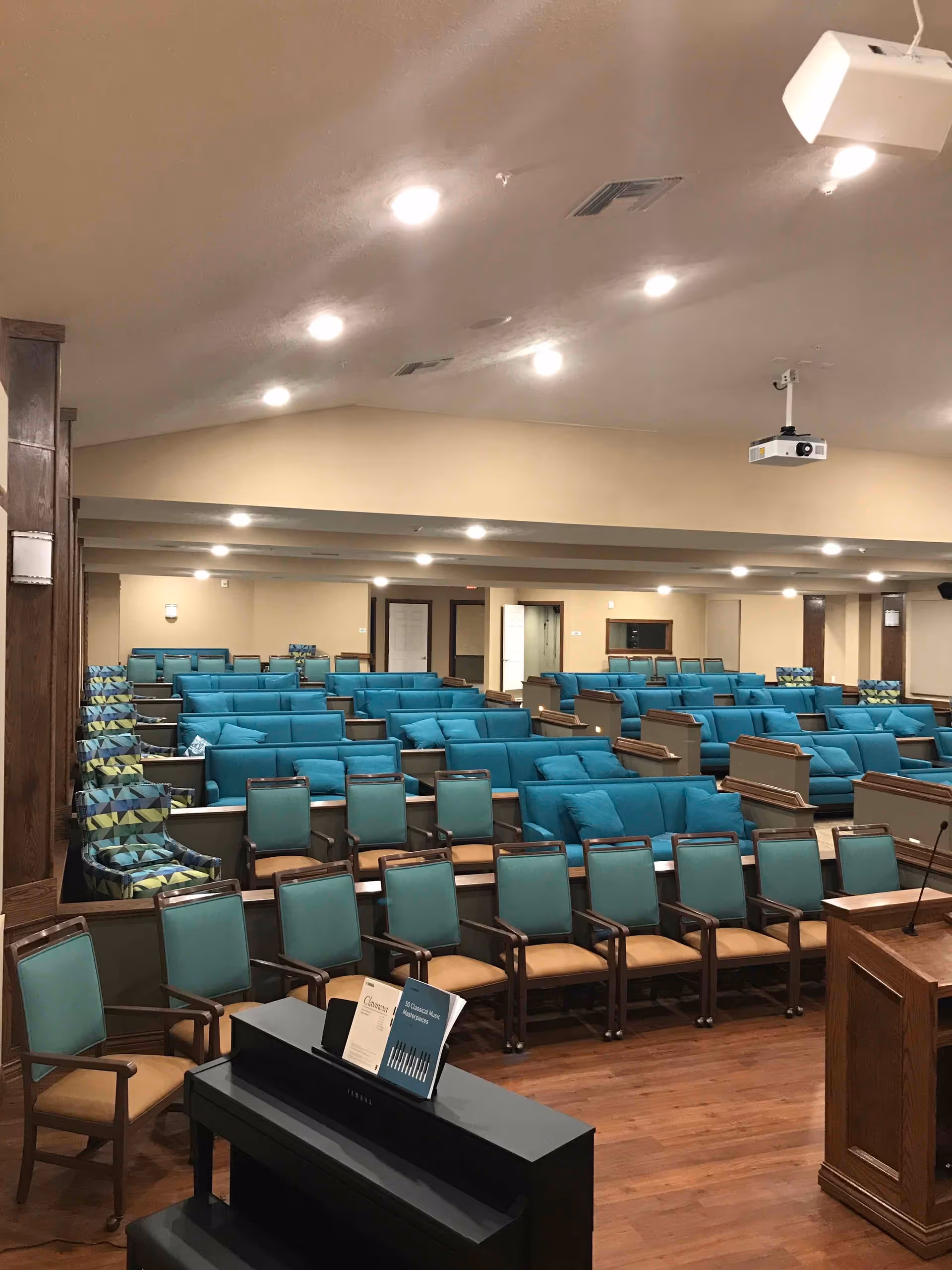 Large interior common room with rows of teal chairs and sofas, a piano and podium, and a ceiling-mounted projector.