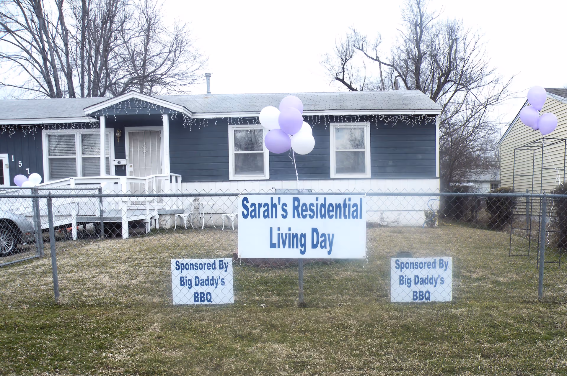 Front yard and facade of a single-story residential home with a sign reading 'Sarah's Residential Living Day' and purple balloons.