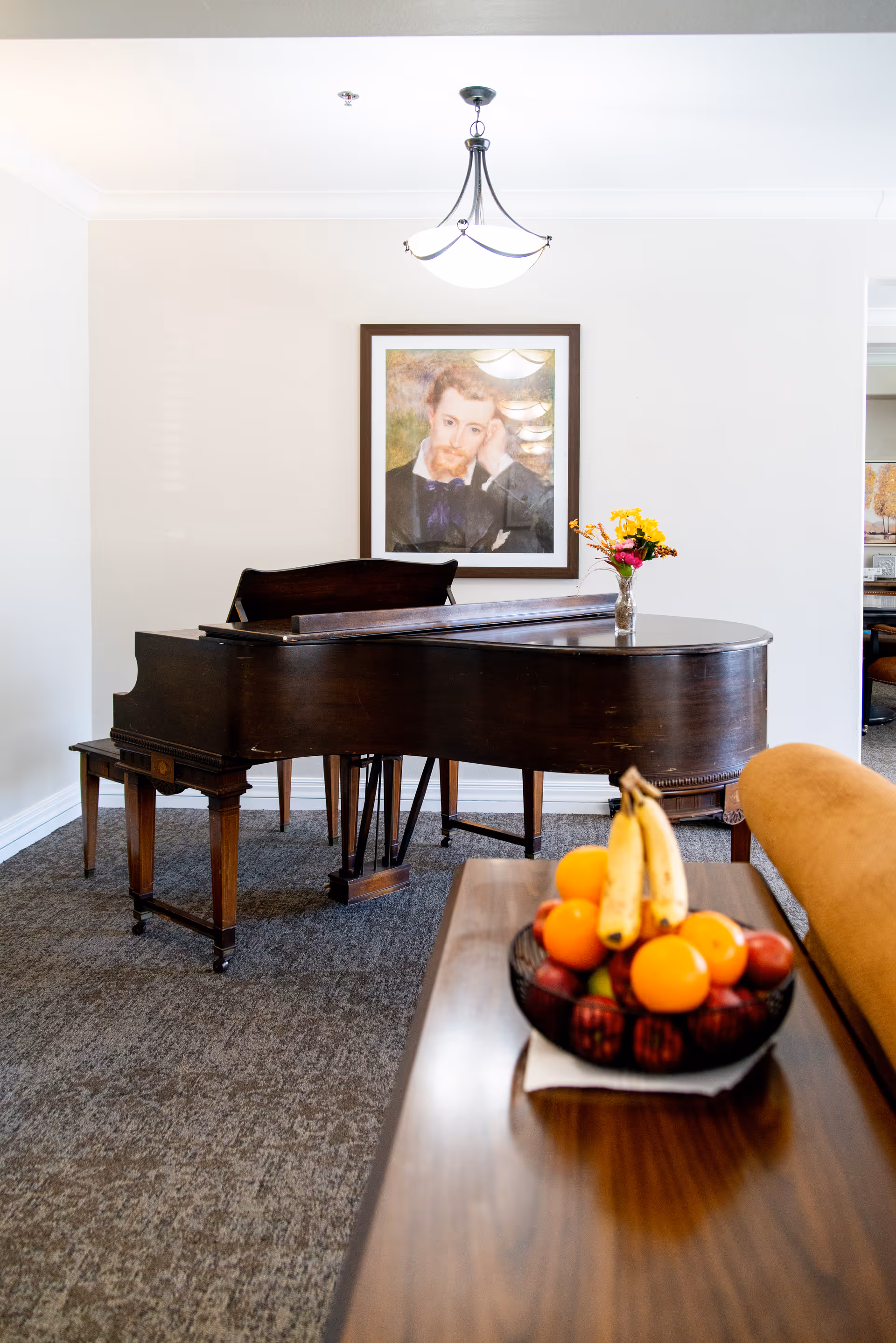 A dark wooden grand piano in a carpeted room with a framed portrait of a man hanging on the wall behind it. A small vase with yellow and pink flowers sits on the piano. In the foreground, there is a wooden table with a bowl of assorted fruits including bananas, oranges, and apples. A mustard-colored chair is partially visible next to the table.