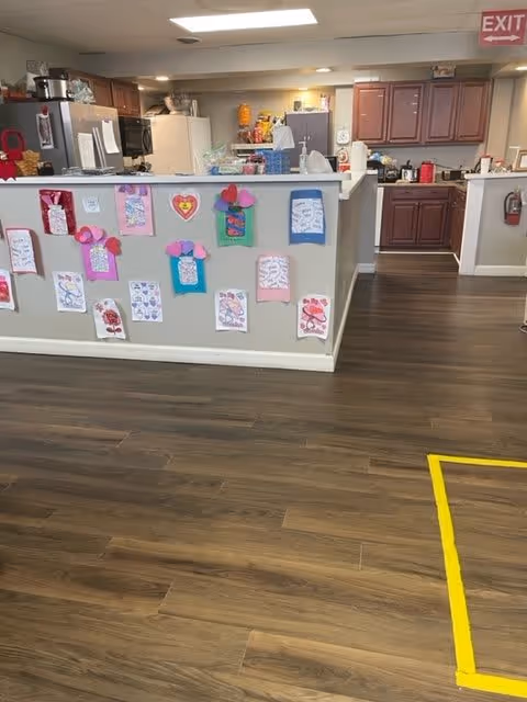 Interior view of a kitchen and common area in a personal care home. The kitchen has wooden cabinets, a refrigerator, microwave, and various kitchen items on the counters. The front side of the kitchen counter is decorated with colorful handmade paper crafts and drawings. The floor is wooden with a yellow tape marking on the right side. An exit sign is visible on the ceiling in the background.