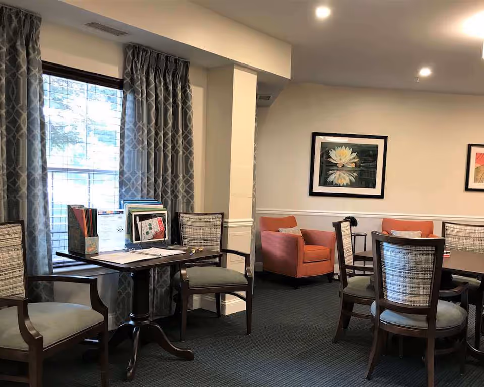 A cozy interior room with a dark wooden table surrounded by cushioned chairs. Two orange armchairs are placed against the far wall, which is decorated with framed floral artwork. A window with patterned curtains lets in natural light, and the room has a calm, inviting atmosphere.