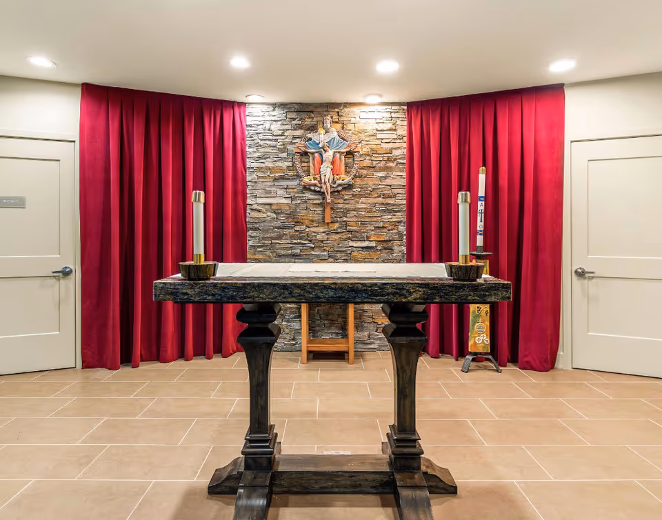 Small chapel interior with an altar table, two candles, red curtains and a religious statue on a stone wall.