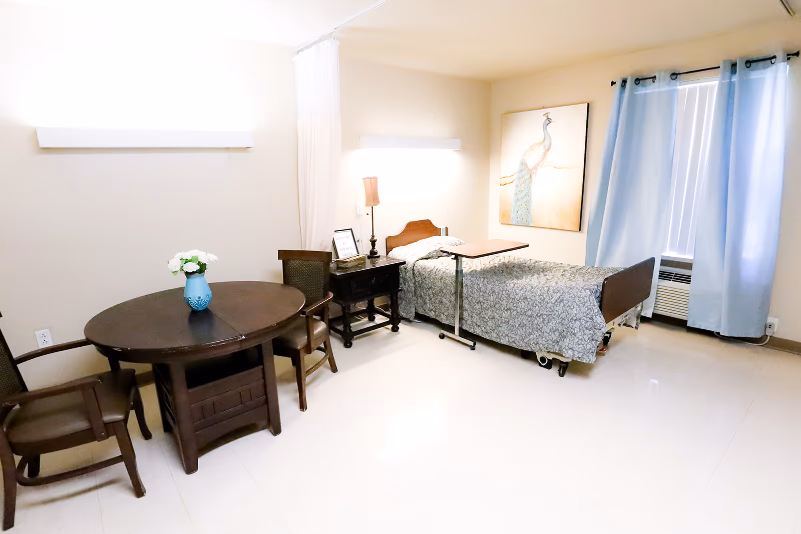 A well-lit room in a healthcare facility featuring a single hospital-style bed with a floral bedspread, a bedside table with a lamp, a round wooden table with a blue vase holding white flowers, and three wooden chairs. The room has light-colored walls, a large window with blue curtains, and a painting of a peacock on the wall.
