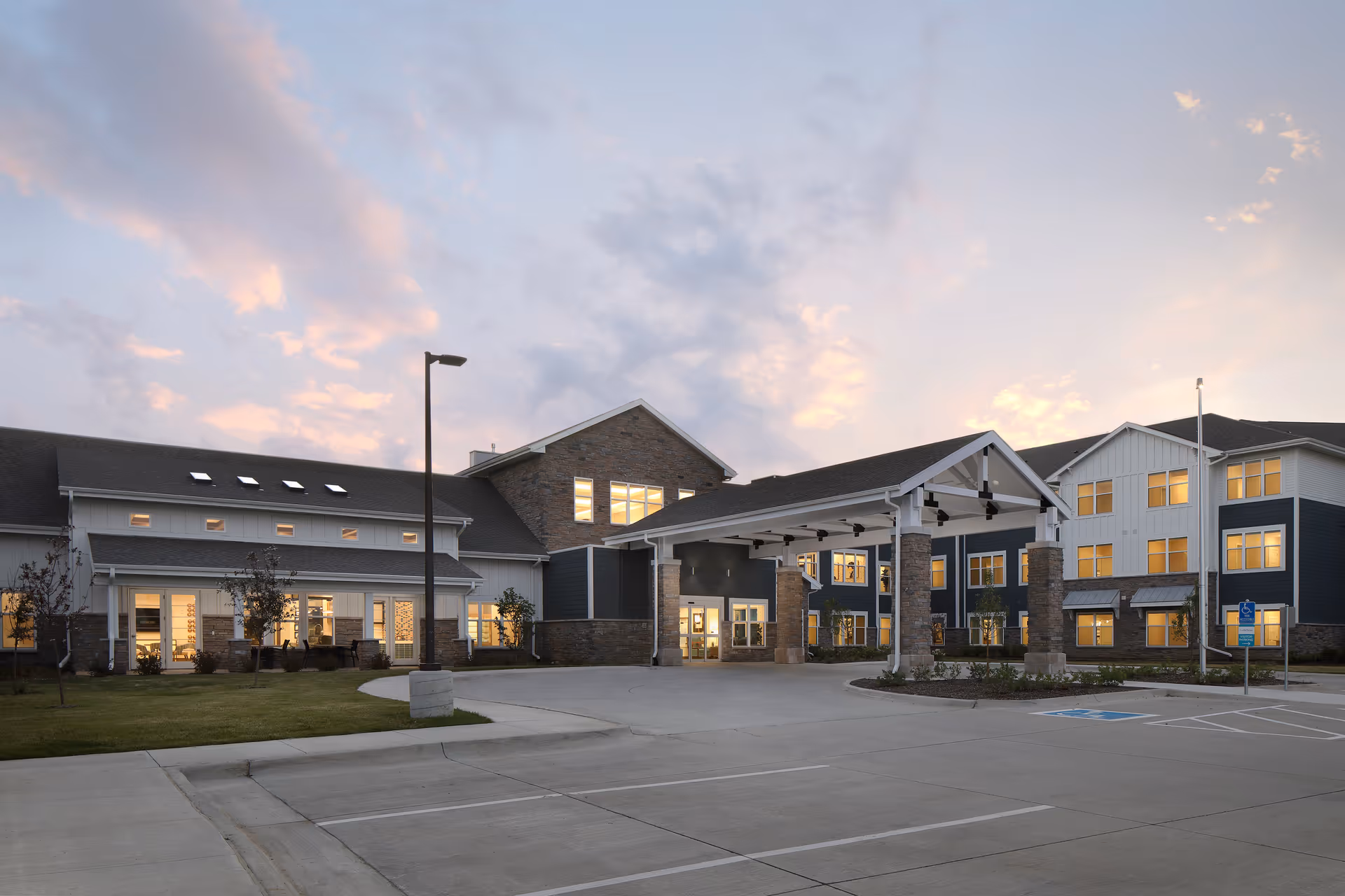 Exterior view of CedarStone Senior Living facility at dusk, showing a large building with multiple windows lit from inside, a covered entrance supported by stone pillars, a parking area with marked spaces, and a partly cloudy sky with soft evening light.