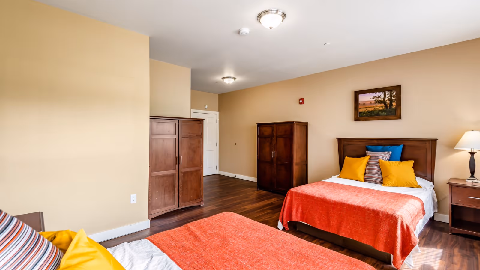 A bedroom with two beds covered in orange blankets and multiple pillows in yellow, blue, and striped patterns. The room has wooden floors, beige walls, two wooden wardrobes, a nightstand with a lamp, and a framed landscape picture hanging on the wall.
