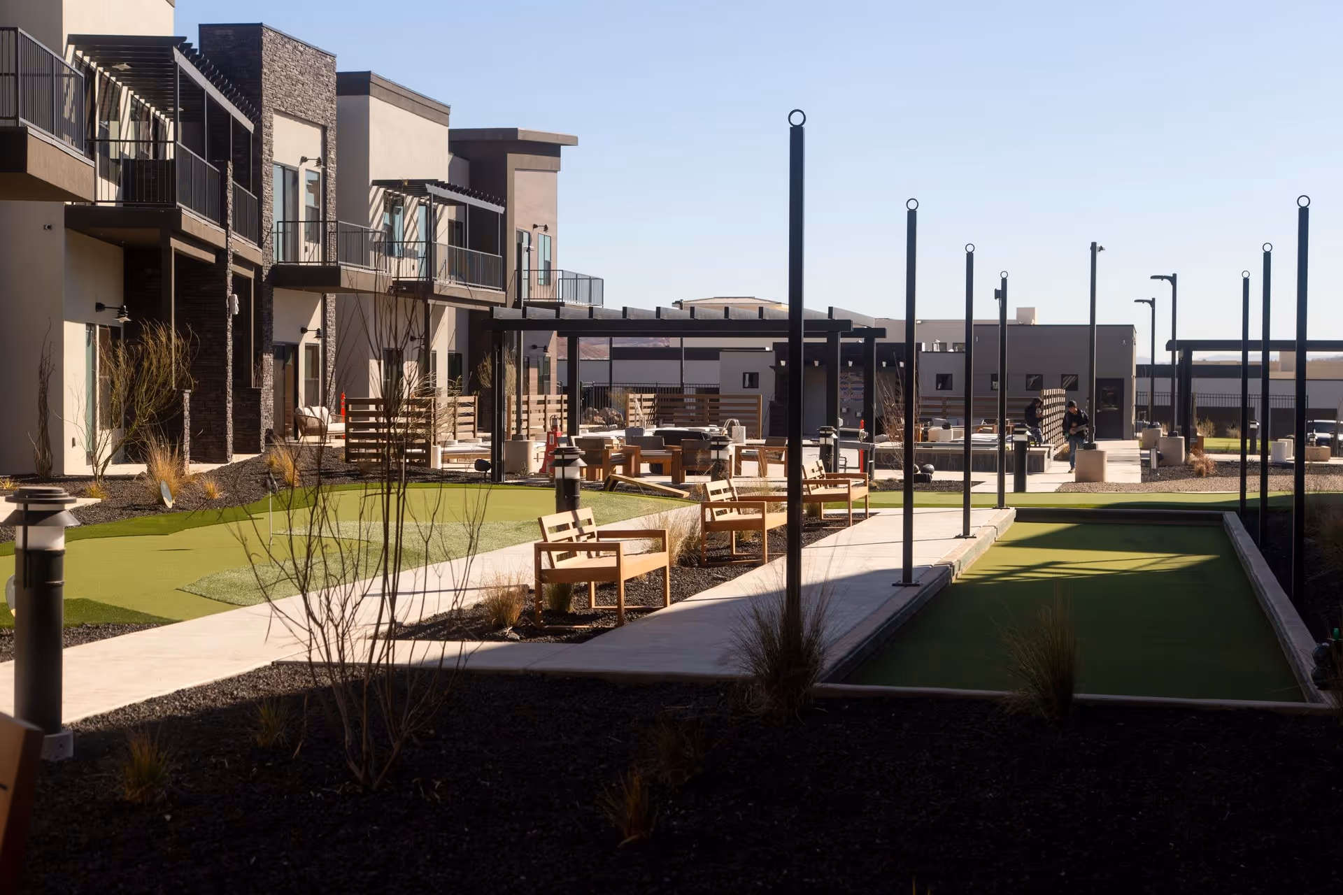 Outdoor courtyard with walkways, benches, artificial turf bocce/green areas and modern two-story apartment buildings in the background.