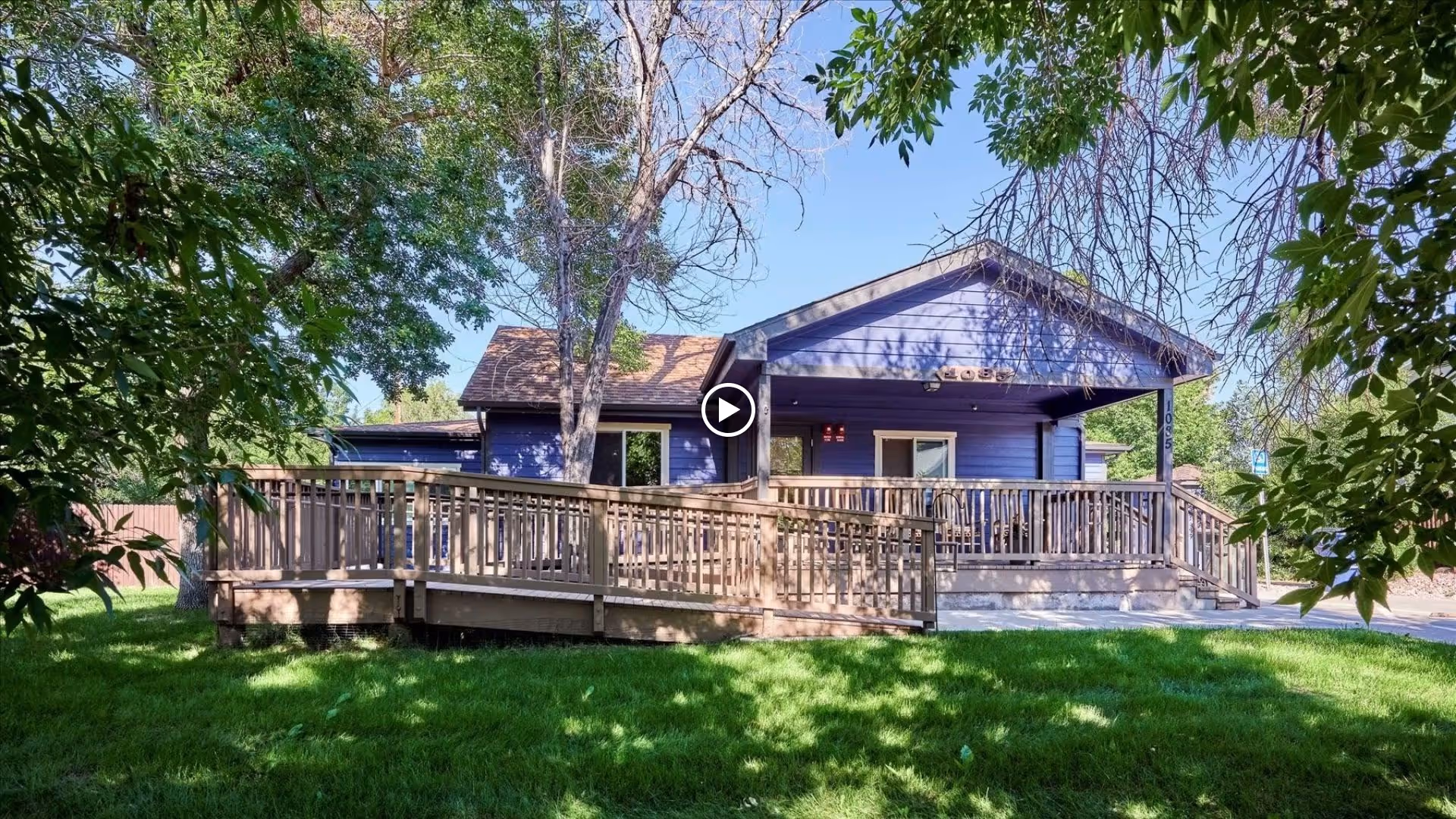 Exterior view of a single-story purple building with a wooden ramp and stairs leading to the entrance, surrounded by green grass and trees under a clear blue sky.