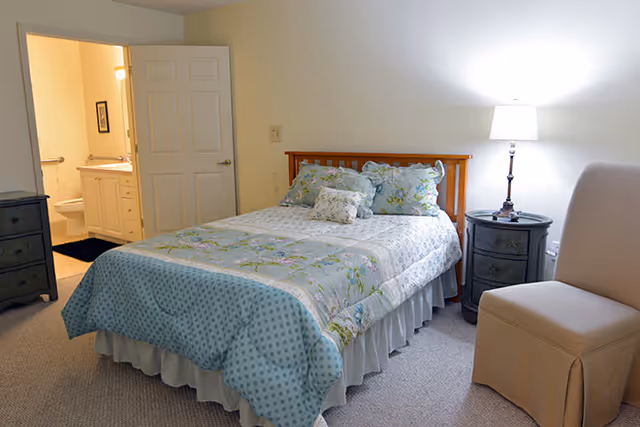 A cozy bedroom with a wooden bed frame, floral and polka dot bedding, and multiple pillows. Next to the bed is a round black nightstand with a lamp on it. A beige upholstered chair is positioned near the nightstand. An open door reveals a bathroom with a sink, mirror, and toilet.
