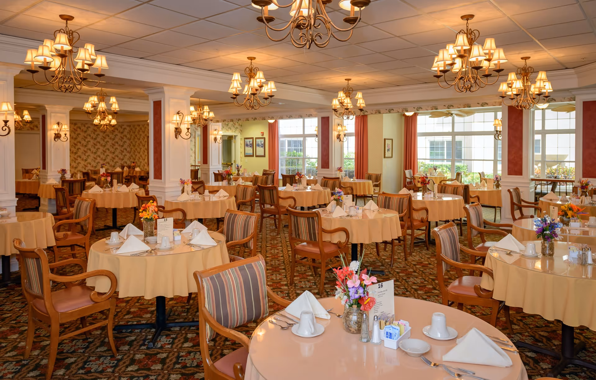 A spacious dining room with round tables covered in beige tablecloths, each set with white napkins, cups, and silverware. The room features wooden chairs with striped cushions, floral centerpieces on each table, ornate chandeliers hanging from the ceiling, and large windows letting in natural light. The carpet has a patterned design and the walls are decorated with wallpaper and framed pictures.