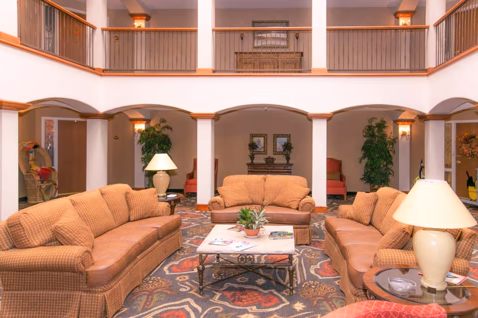A spacious and well-lit living room area with three brown upholstered sofas arranged around a rectangular coffee table with a plant and magazines on it. The room features a patterned carpet, two table lamps on side tables, potted plants, and a second-floor balcony with railings overlooking the area. The walls have arches and decorative lighting fixtures.