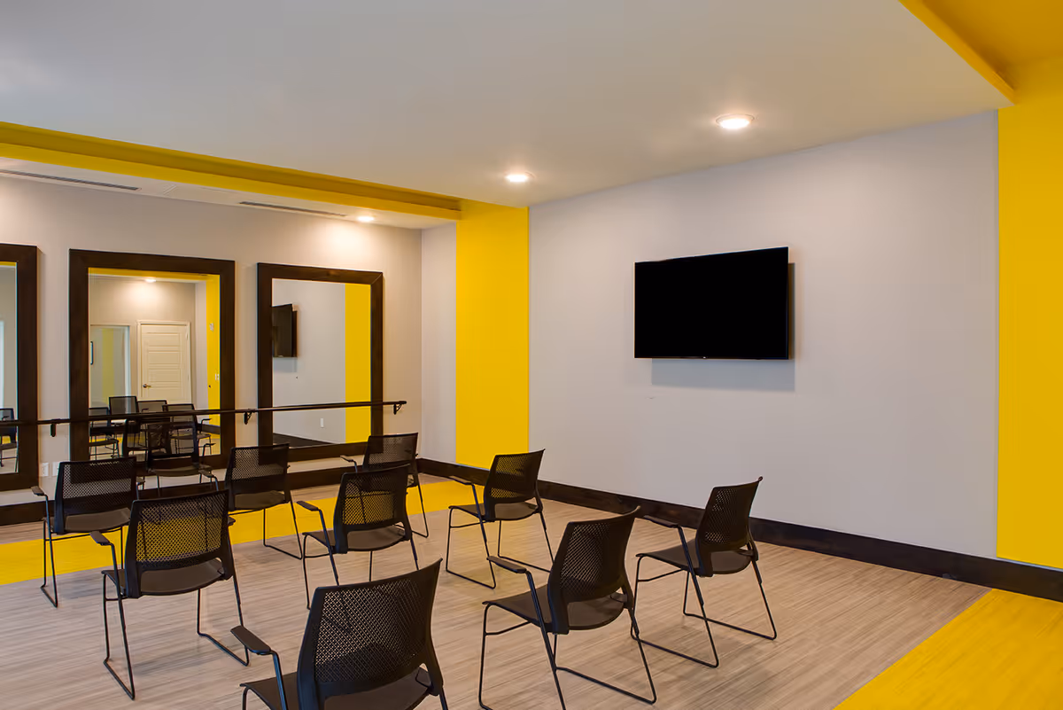 A small meeting or exercise room with black mesh chairs arranged in rows facing a wall-mounted flat screen TV. The room features bright yellow accents on the walls and ceiling, large mirrors on one side, and light wood flooring.