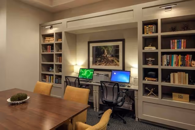 A cozy interior room featuring a wooden table with four upholstered chairs in the foreground. In the background, there are two computer workstations with black mesh chairs, a printer, and built-in bookshelves filled with books and decorative items. A framed picture of tree roots hangs on the wall between the workstations, and two small lamps provide warm lighting.