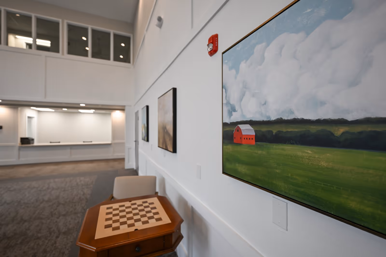 Interior view of a senior living facility hallway with a chess table and chair in the foreground. The walls are decorated with paintings, including one of a red barn in a green field under a cloudy sky. The area is well-lit with overhead lights and has a carpeted floor.