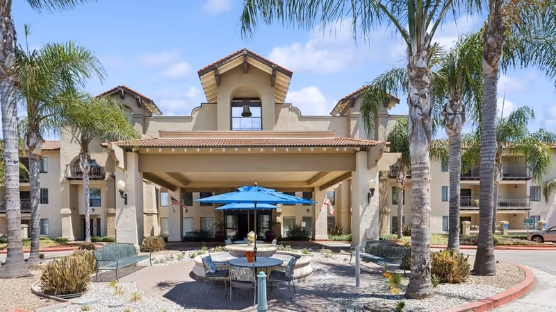 Exterior view of a senior living facility named Rio Las Palmas featuring a beige stucco building with a covered entrance, palm trees, outdoor seating with a round table and blue umbrella, and a clear blue sky.
