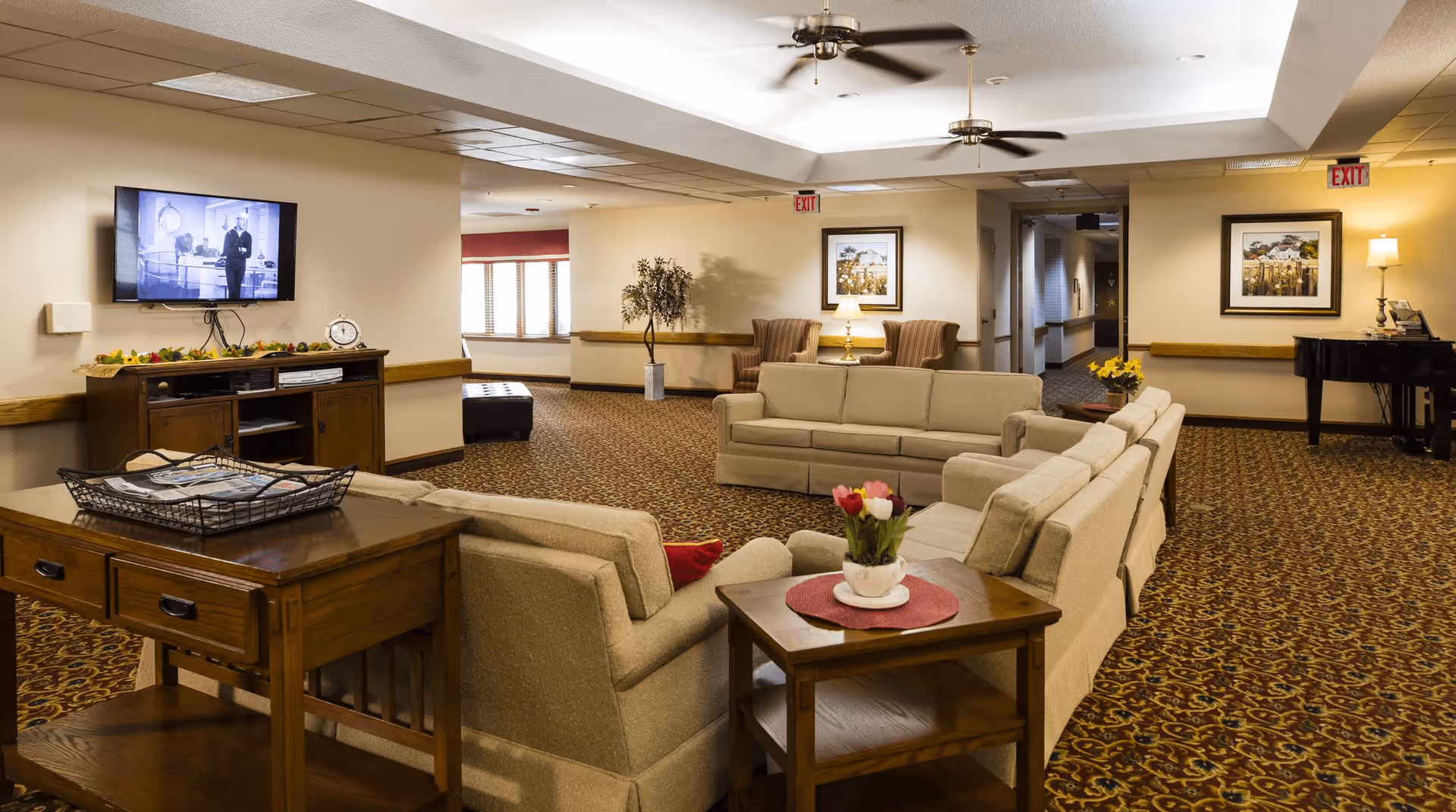 Spacious assisted-living common area with beige sofas arranged around side tables, a wall-mounted TV, decorative lamps and a piano.