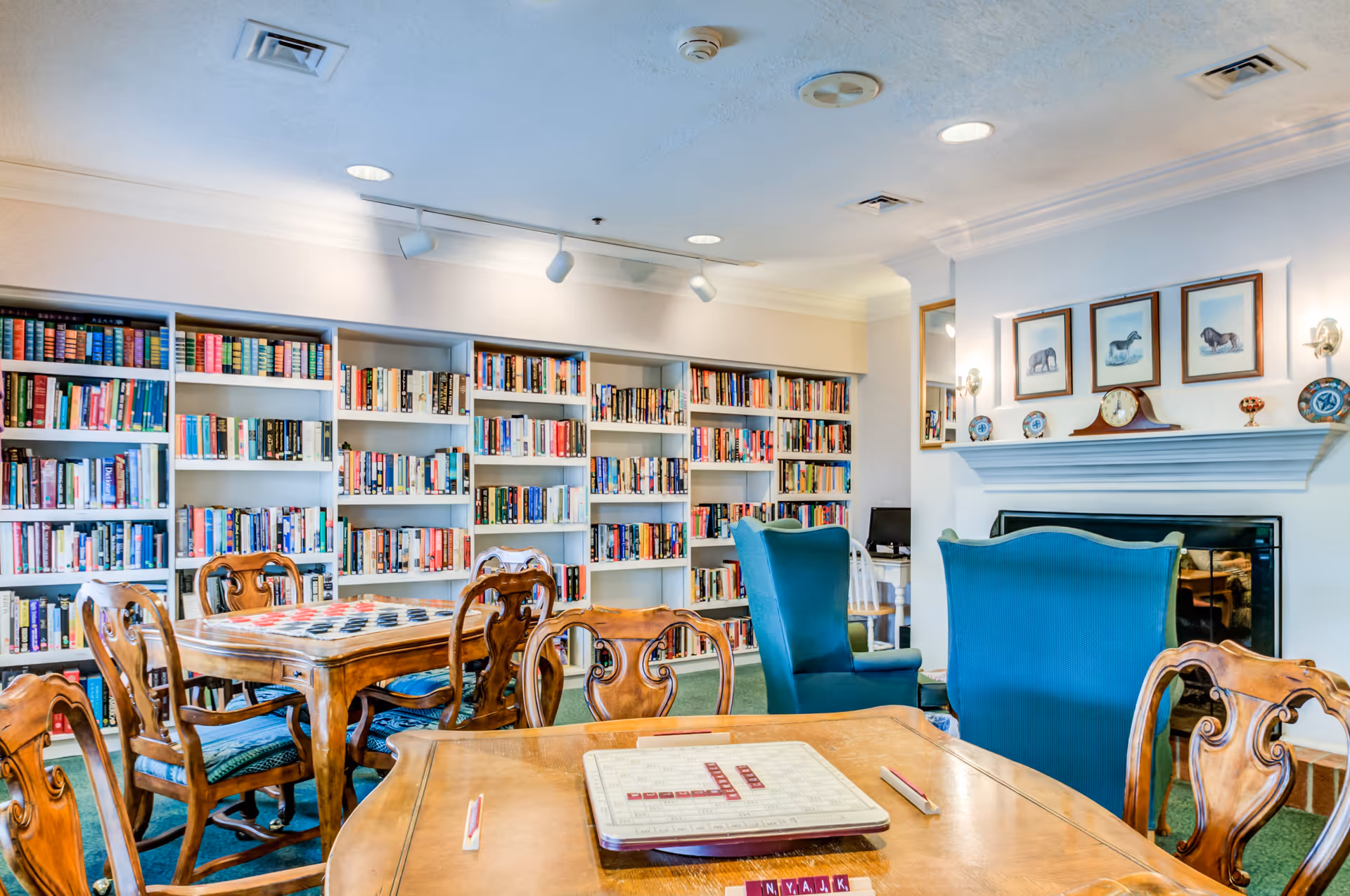A cozy library room with wooden tables and chairs, two blue armchairs facing a fireplace with framed animal pictures above it, and bookshelves filled with books along the back wall. The room has a warm and inviting atmosphere.