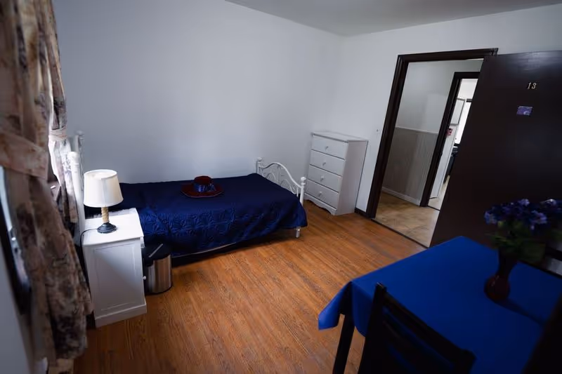 Simple senior bedroom with a single bed, nightstand and lamp, dresser, small table with a blue tablecloth, and wood-look flooring.