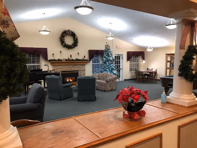 A decorated senior living common room with chairs around a fireplace, a Christmas tree, and a poinsettia on a wooden counter.