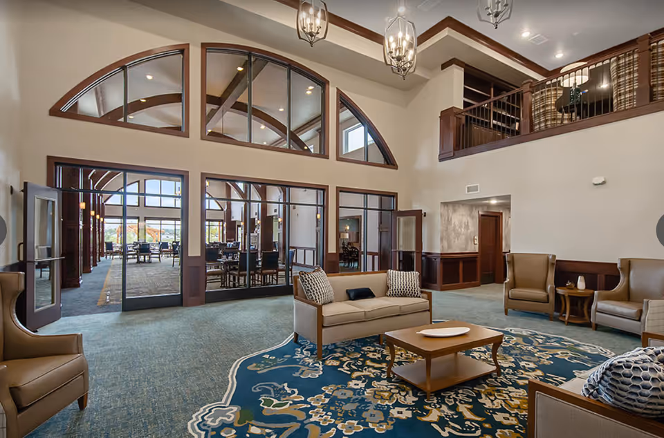 Spacious senior living facility common area with high ceilings, large arched windows, and glass doors leading to a dining area. The room features comfortable seating including sofas and armchairs arranged around a wooden coffee table on a decorative blue and beige rug. There is a balcony with railing overlooking the space and elegant hanging light fixtures.