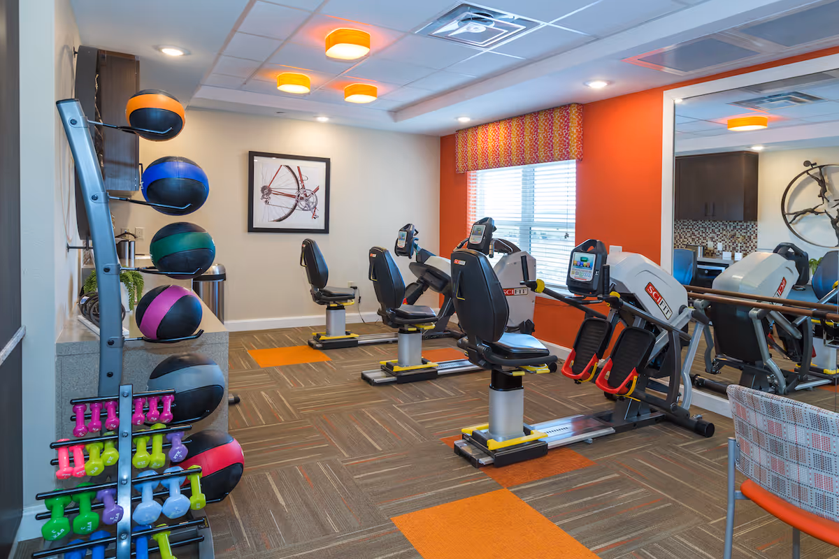 A small fitness room with exercise equipment including three recumbent exercise bikes, a rowing machine, a rack of colorful medicine balls, and various dumbbells. The room has a large mirror on one wall, a window with blinds and a patterned valance, and bright orange accent walls. The floor is carpeted with a pattern that includes orange squares.