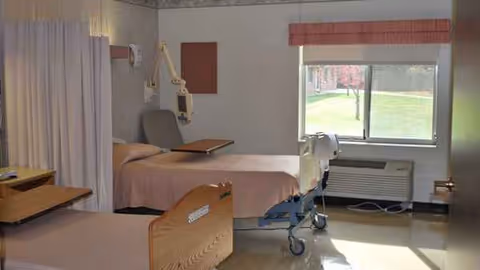A hospital-style patient room with two beds, privacy curtains, medical equipment, and a window.