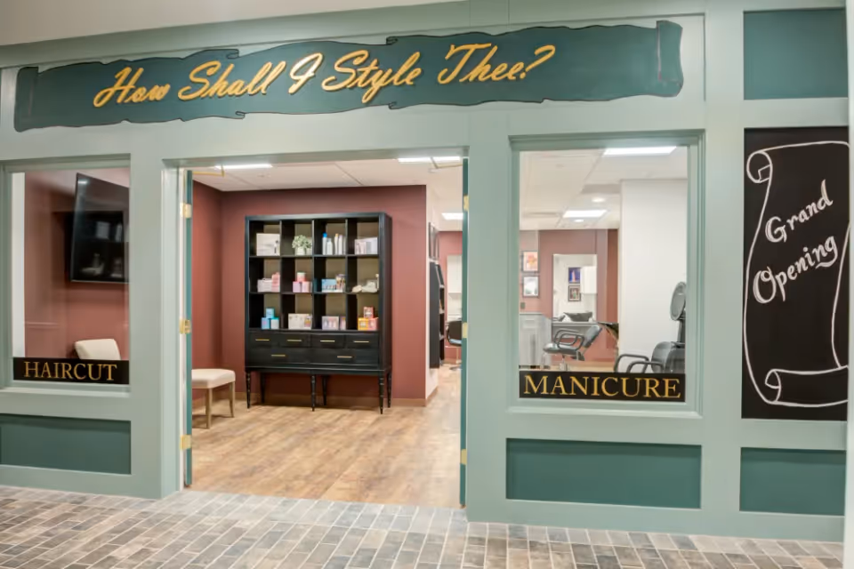 Interior view of a salon area in a senior living facility with a sign above the entrance reading 'How Shall I Style Thee?'. The entrance has two windows labeled 'HAIRCUT' and 'MANICURE'. Inside, there are chairs, a black shelving unit with various hair and beauty products, and salon stations visible in the background. A chalkboard sign on the right side says 'Grand Opening'.