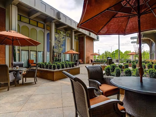 Outdoor courtyard patio with round tables, wicker chairs, and orange umbrellas beside a brick building.