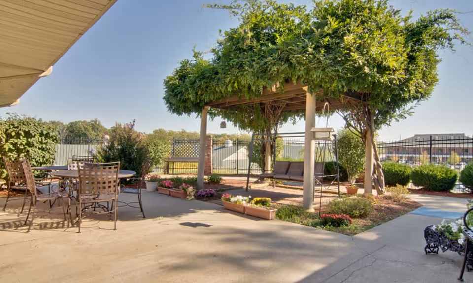 Outdoor patio area with a round metal table and chairs on the left, a pergola covered with green vines and a hanging swing bench in the center, surrounded by flower beds and shrubs under a clear blue sky.