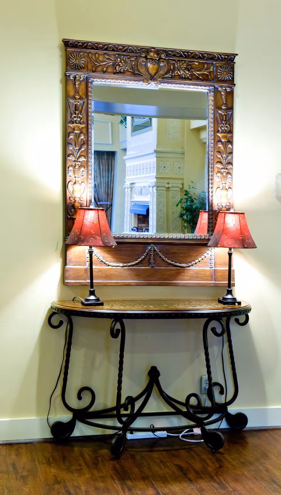 An ornate wooden-framed mirror mounted on a pale yellow wall above a decorative wrought iron and wood console table. Two red lamps with black bases are placed symmetrically on the table, casting warm light. The mirror reflects part of an elegant interior with detailed molding and a plant.