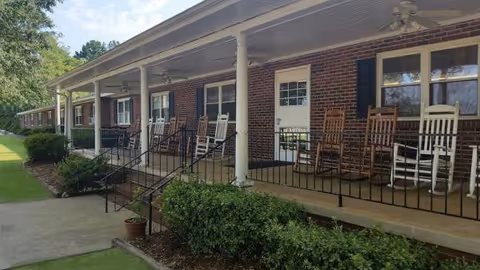 Brick facility exterior with a long porch lined with rocking chairs and greenery