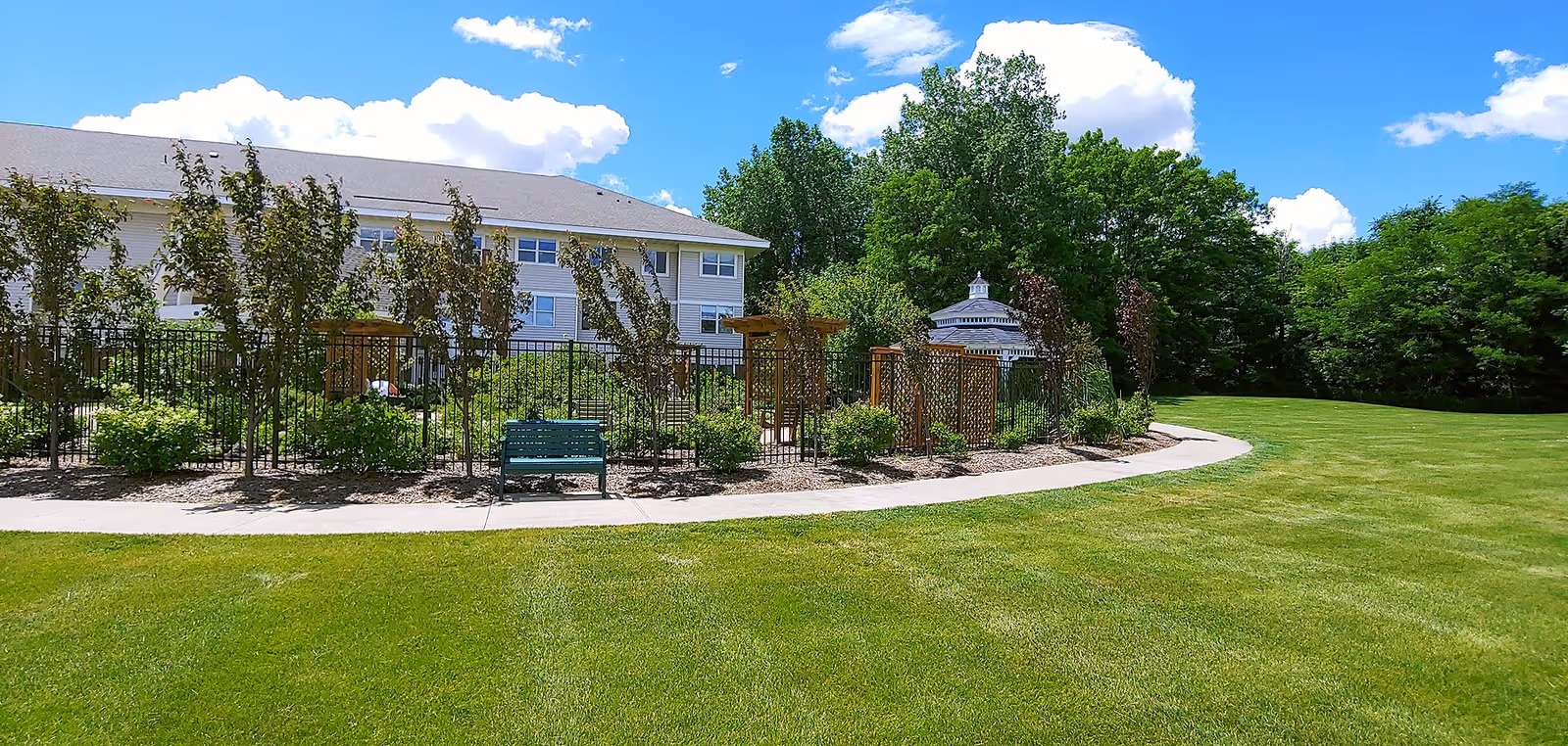 A well-maintained outdoor garden area at Oak Meadows Senior Living featuring a green lawn, a paved walkway, a green bench, a fenced garden with small trees and shrubs, and a white gazebo in the background under a blue sky with scattered clouds.